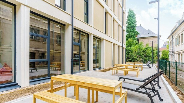 An outdoor patio area with wooden tables and benches, large windows, and a green fence. - Student Factory Nancy Centre (Nancy)