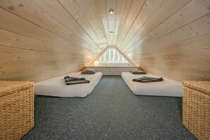 A room with a sloped ceiling, two white beds with pillows, a woven bench, and a view through a window.