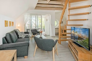 A modern living room with a sofa, a TV on a wooden stand, a staircase, and a view through large windows.