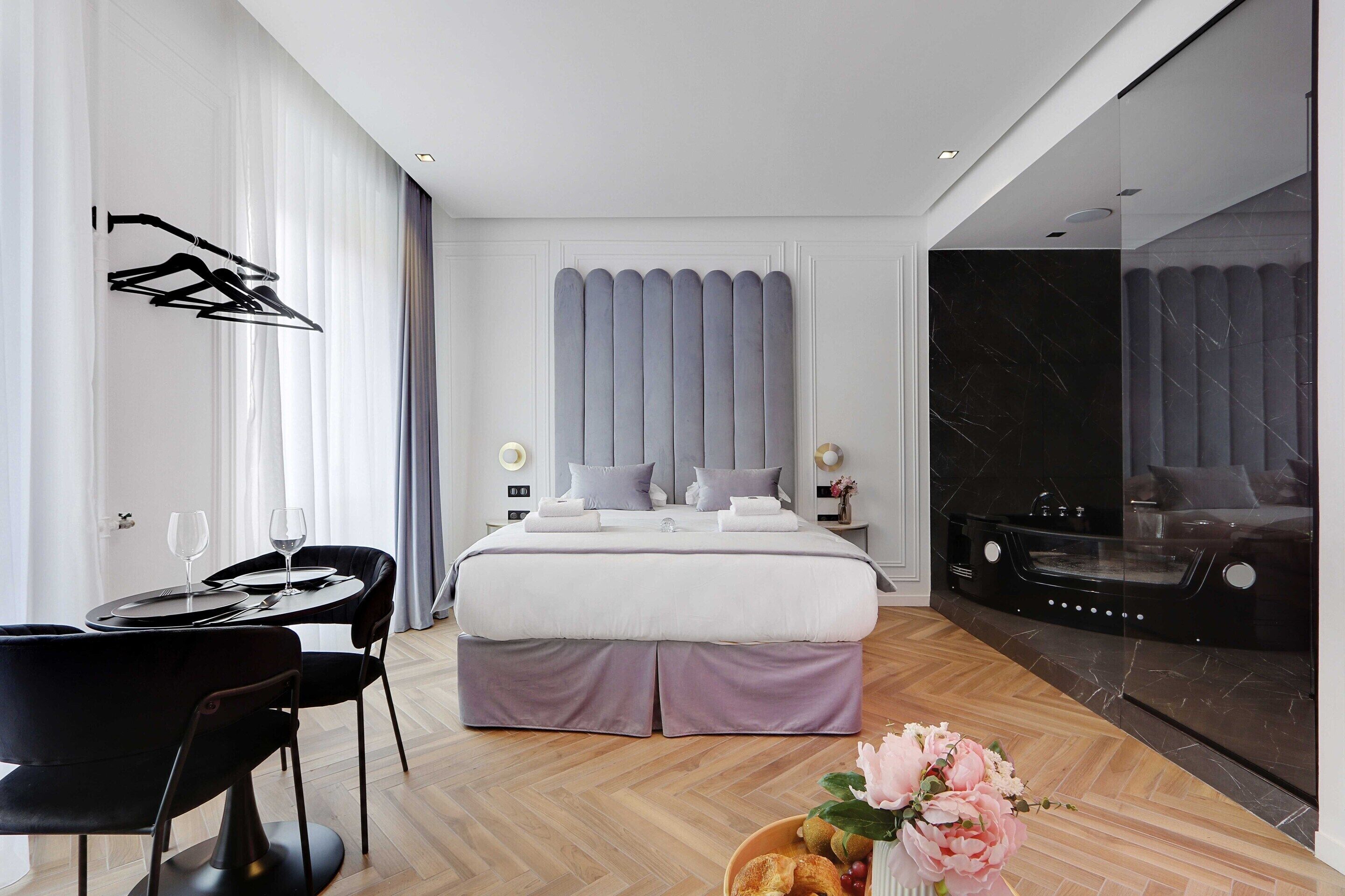 Een moderne hotelkamer met een netjes opgemaakt bed, een zithoek met een tafel en stoelen, een badkamer met een bad, en een grote spiegel die de kamer weerspiegelt.