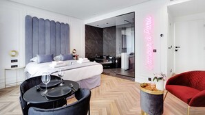 Une chambre d’hôtel moderne avec un lit, une table à manger avec des chaises, un fauteuil rouge, une petite table avec un vase de fleurs et un néon mural.