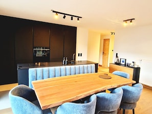 Cozinha moderna com armários escuros, uma ilha central e um espaço de jantar com mesa de madeira e cadeiras estofadas em azul.