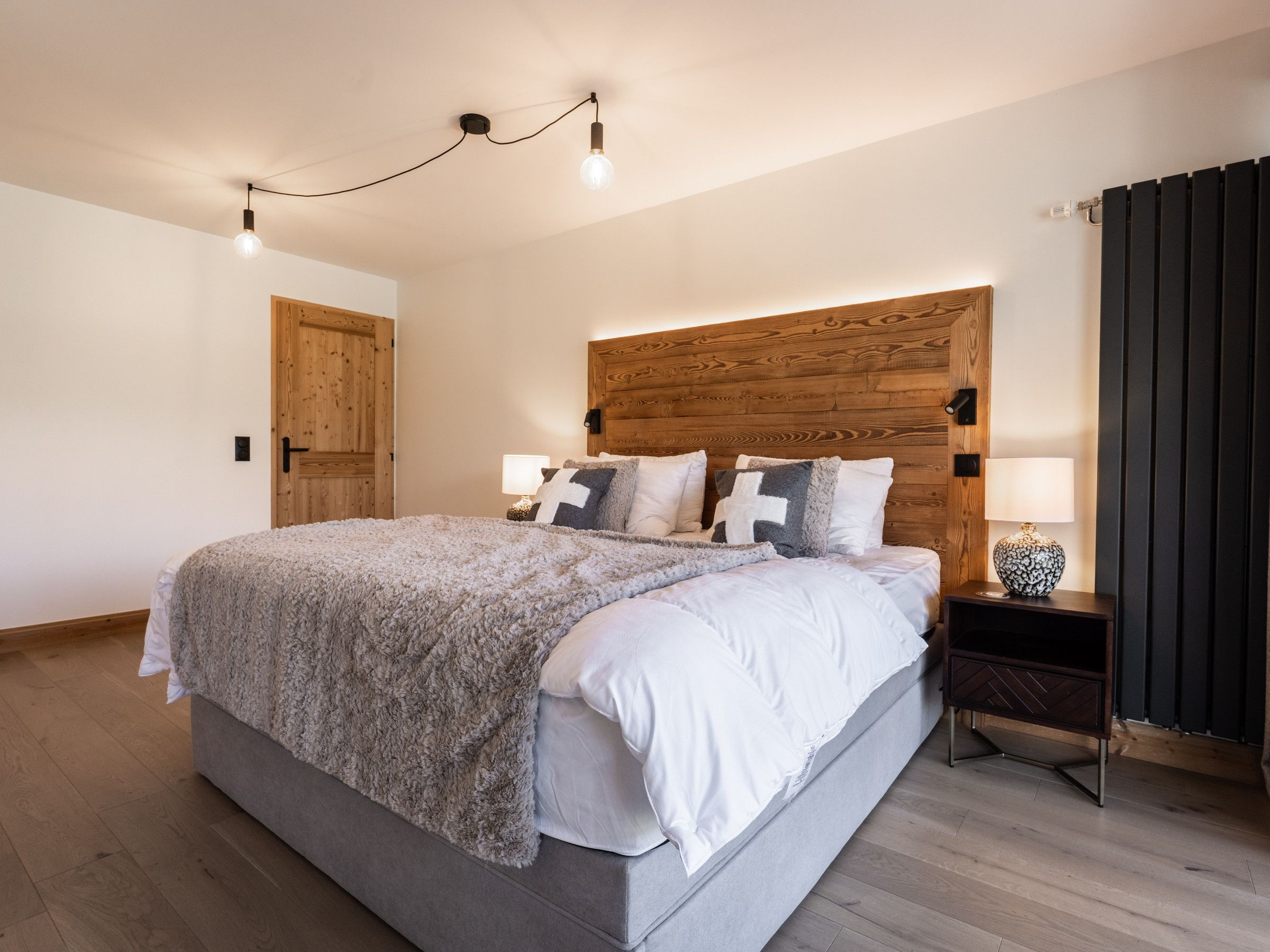 A bedroom with a wooden headboard, a bed with white and gray bedding, a nightstand with a lamp, and a wooden door.