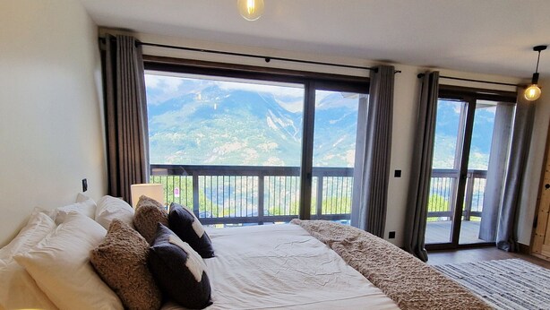 A bedroom with a large bed, a nightstand, a lamp, and a view of mountains through the windows.