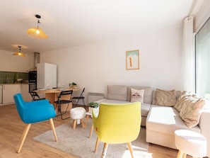 A modern living room with a sofa, two chairs, a dining table with chairs, a kitchen area, and a large window.