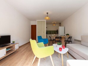 A compact living space with a TV, a sofa, two chairs, a dining table, and a kitchen area.