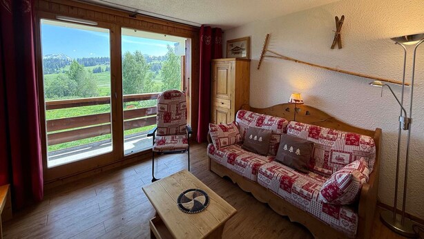 Una sala de estar acogedora con suelo de madera, un sofá estampado, una mesita y vistas a un paisaje verde a través de amplios ventanales.