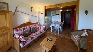 Una sala de estar acogedora con un sofá, una mesa de madera y un sillón de mimbre. Se ve una cocina al fondo con una mesa y sillas de comedor.