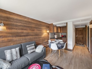 Een moderne woonkamer met een grijze bank, een eethoek met een tafel en stoelen, houten wanden en een keuken op de achtergrond.