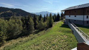 A building with a stone base and wooden upper levels, set against a backdrop of a forested hillside and distant mountains. - New 3-room apartment, 6 guests, terrace, parking, Les Saisies (Villard-sur-Doron)