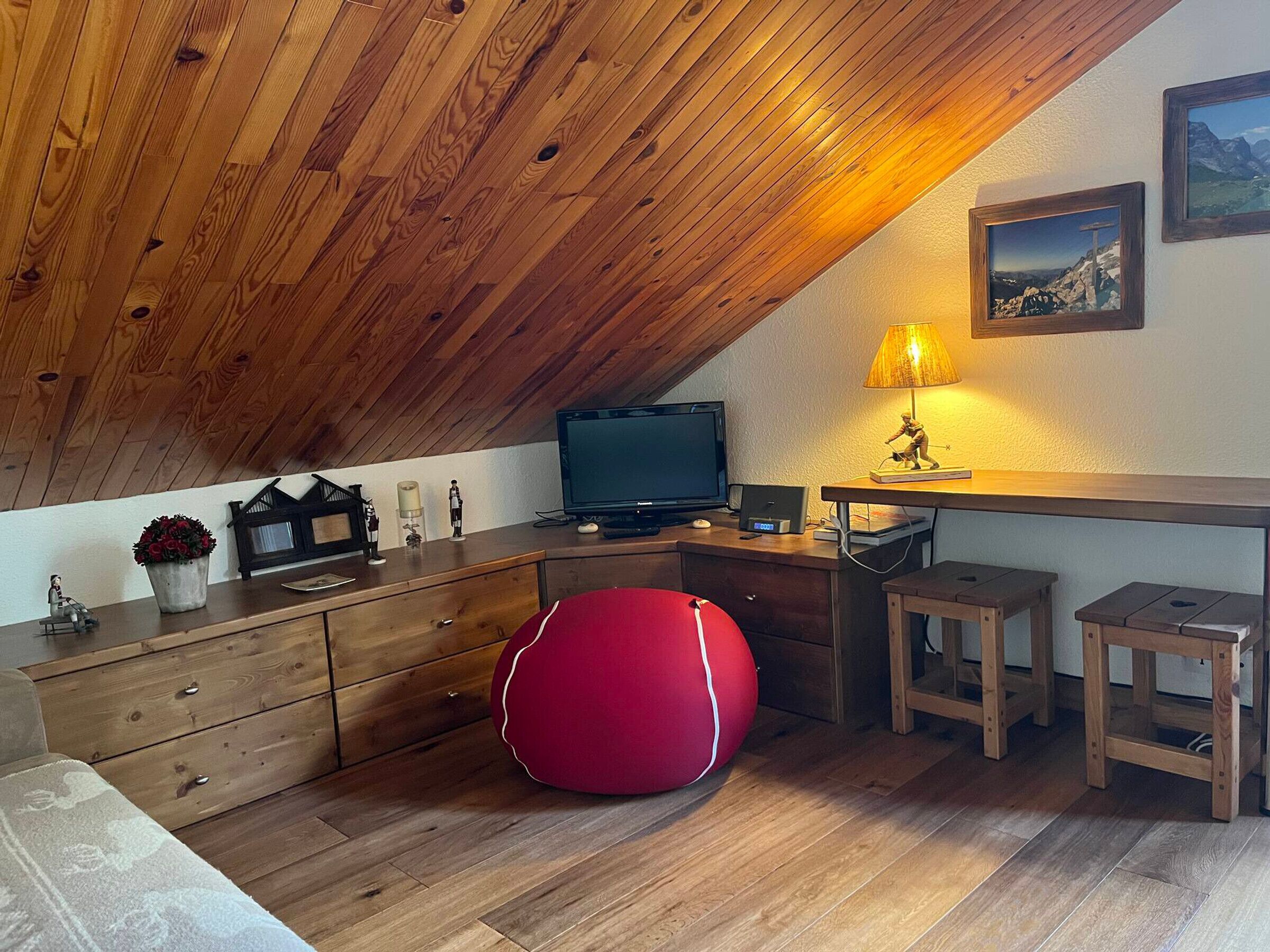 Acogedor dormitorio en el ático con vigas de madera, televisor, escritorio con lámpara y un sillón de peluche rojo.