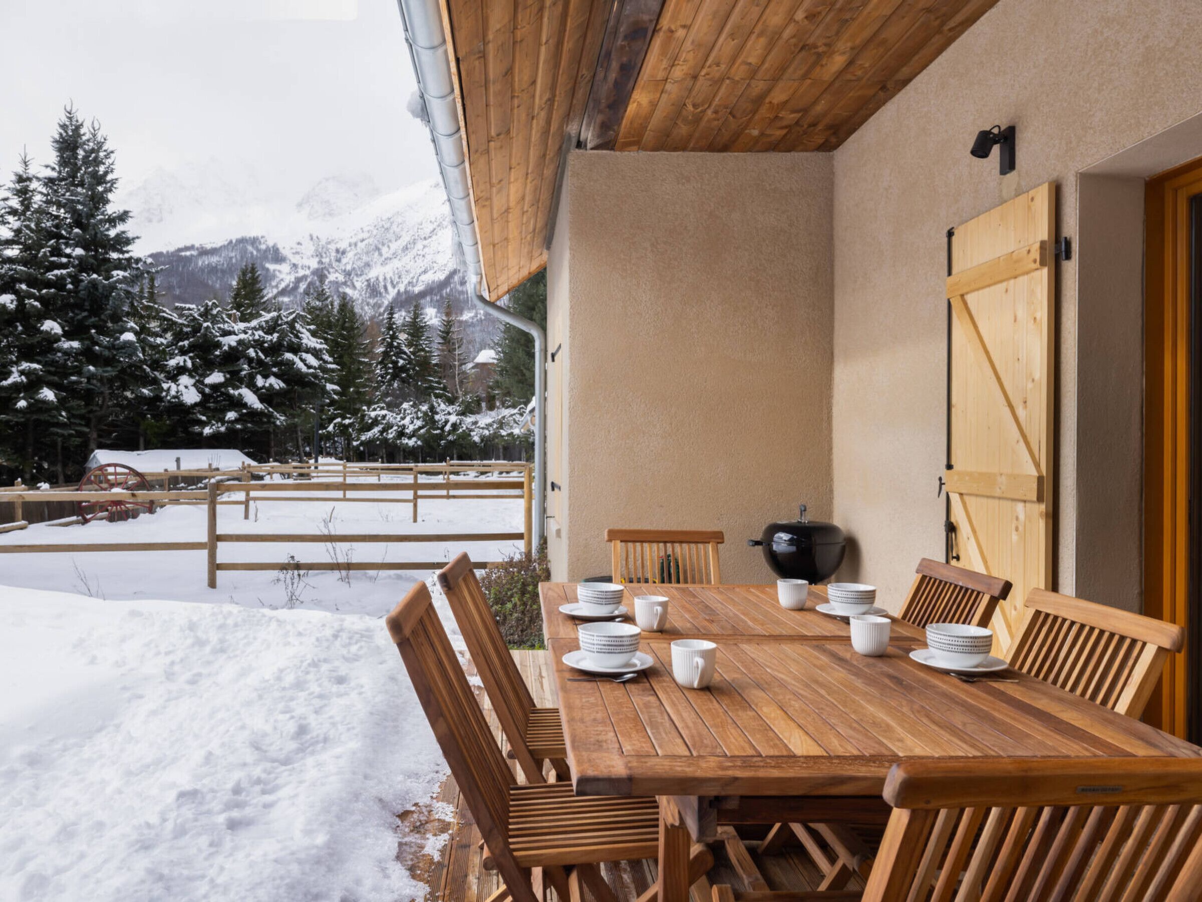 A wooden dining table set for six with chairs, a black grill, and a view of snow-covered trees and mountains.