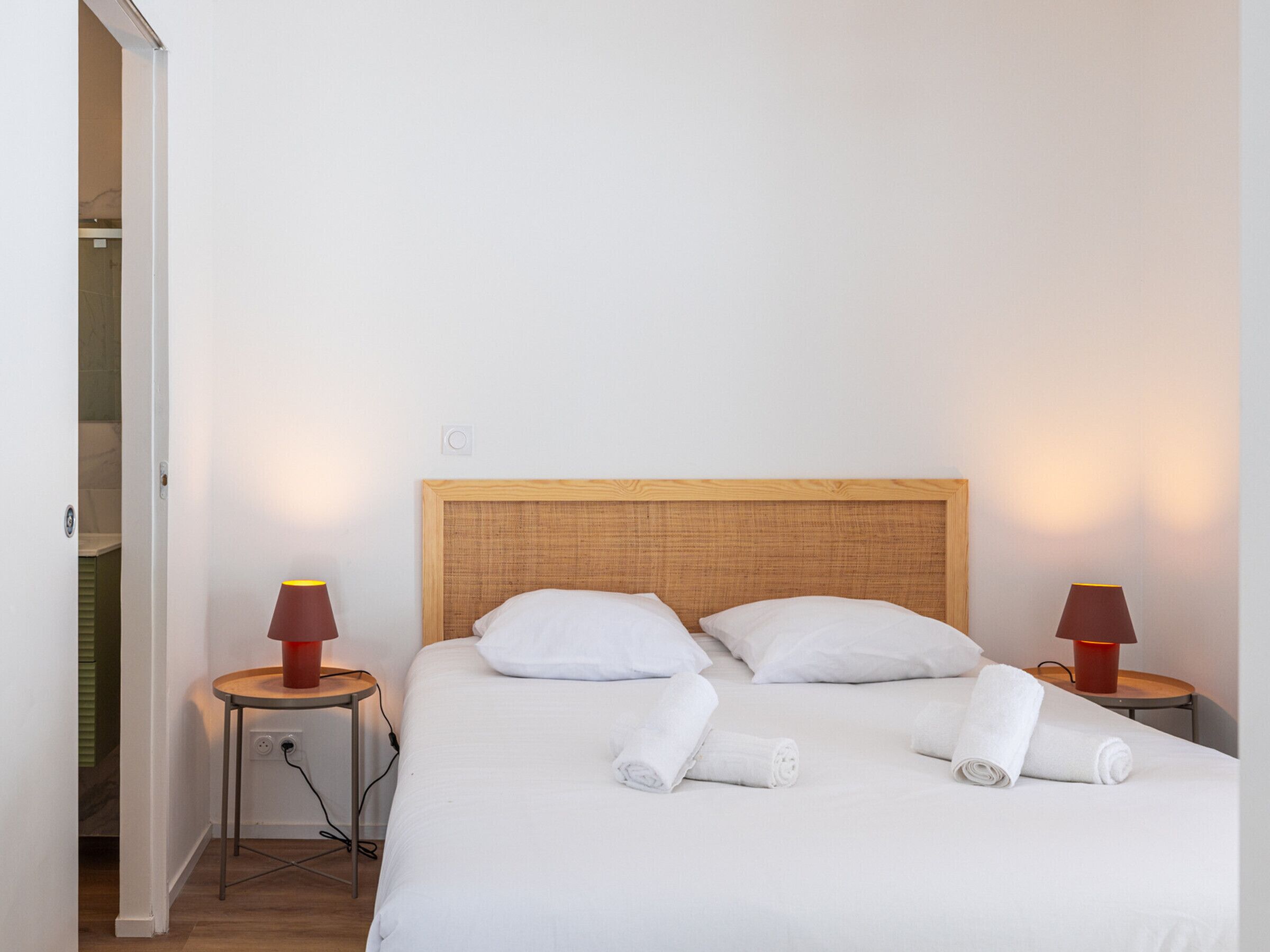 Ein ordentlich bezogenes Bett mit weißen Leinen und zwei aufgerollten Handtüchern, ein Holz-Kopfteil und zwei Nachttischlampen mit roten Lampenschirmen.