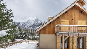 A wooden chalet with a balcony, surrounded by snow-covered trees and mountains in the background. - New Apartment with Garden and Terrace near the Village (Le Monêtier-les-Bains)