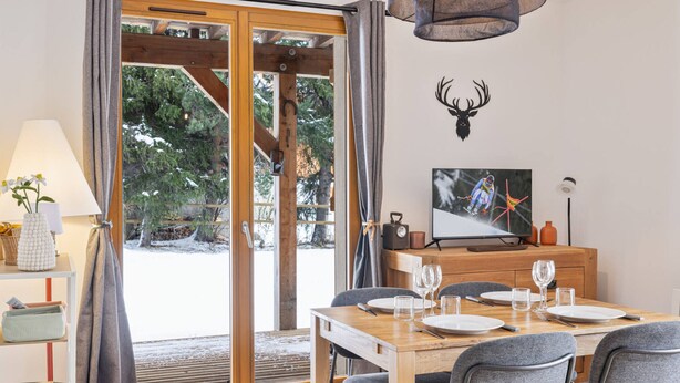 A dining area with a wooden table set for four, a television mounted on the wall, and a door leading to an outdoor area with a view of trees and snow. - New Apartment with Garden and Terrace near the Village (Le Monêtier-les-Bains)