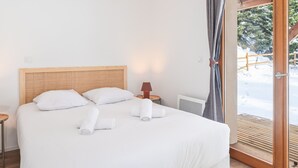 A bedroom with a wooden headboard, a bed with white linens, two rolled towels, a bedside lamp, and a view of a snowy outdoor area through a glass door. - New Apartment with Garden and Terrace near the Village (Le Monêtier-les-Bains)