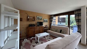A living room with a sofa, a coffee table, a TV, and a wooden cabinet. There is a window with a view of the outside.