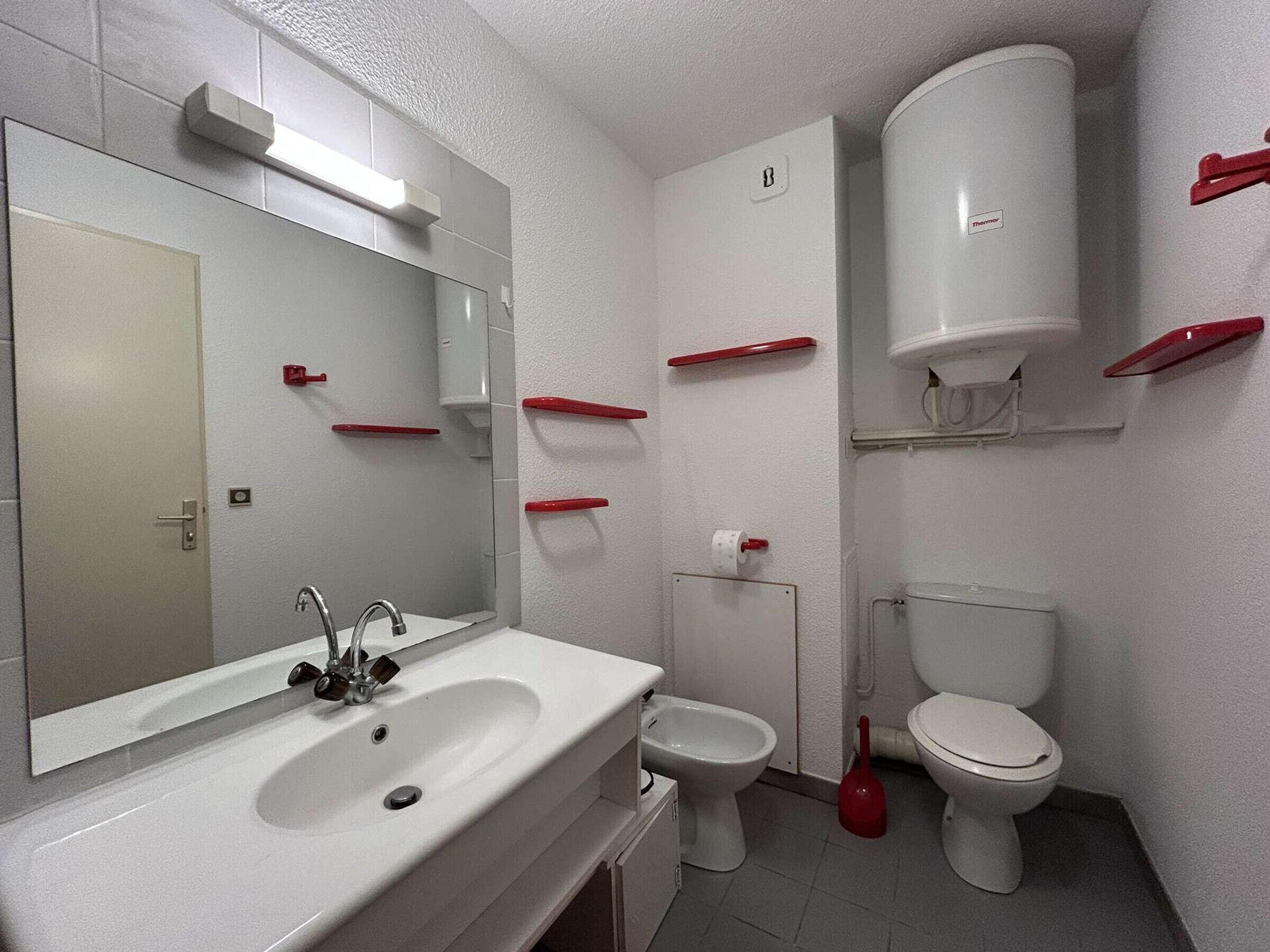 Une salle de bain avec un lavabo, deux toilettes, un miroir et une ventouse rouge.