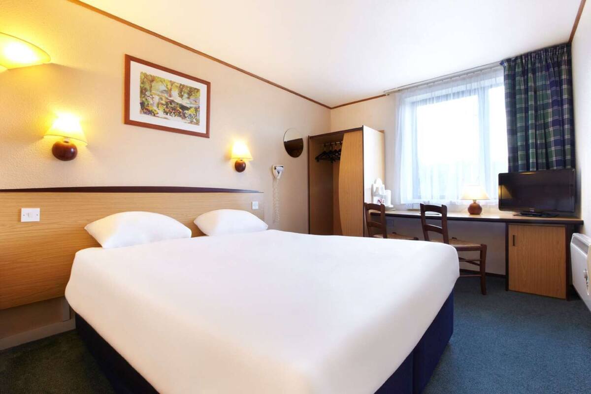 A hotel room with a large bed, a desk with a computer and chair, a television, and a window with curtains.