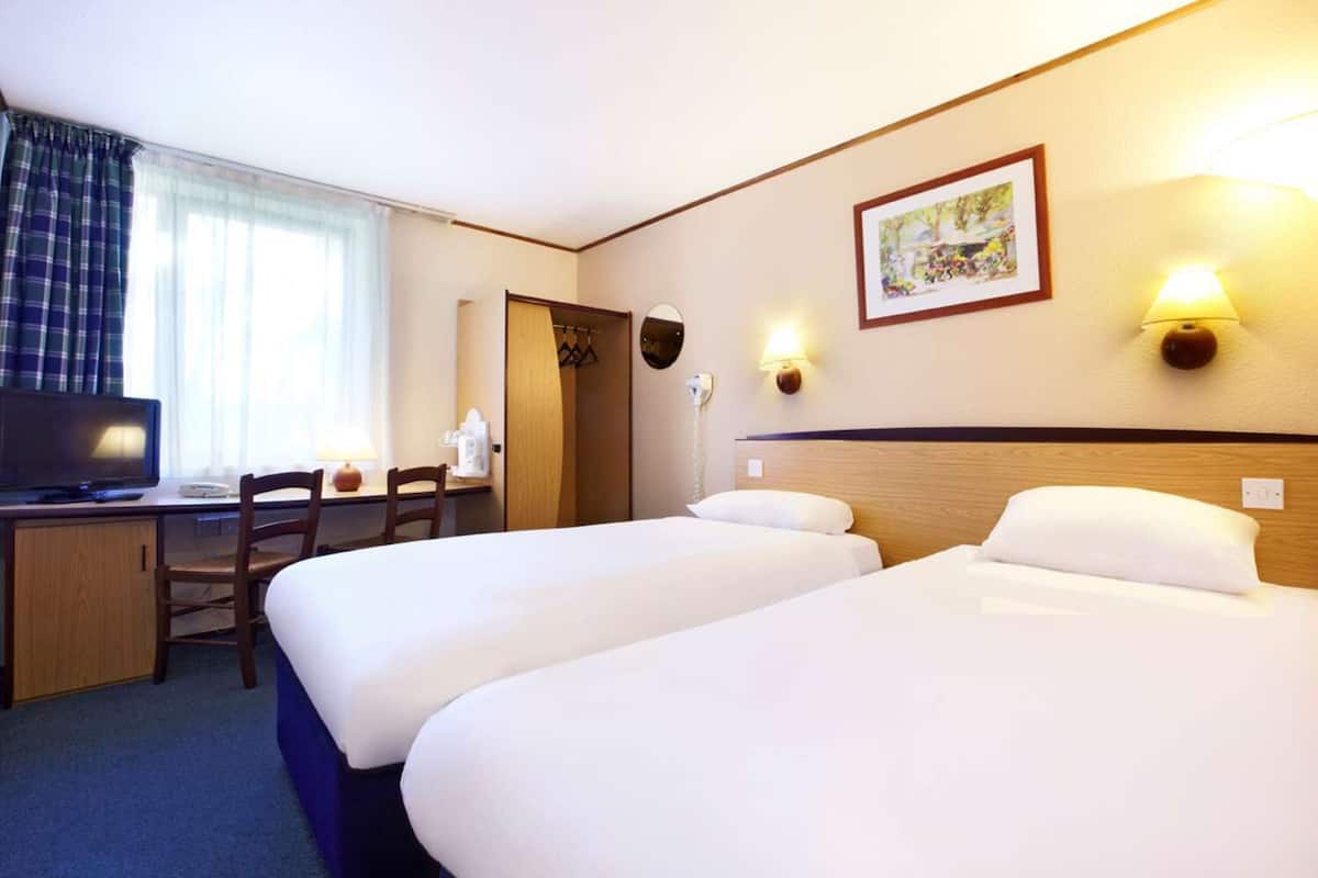 A hotel room with two beds, a desk with a computer, a chair, a TV, and a window with curtains.