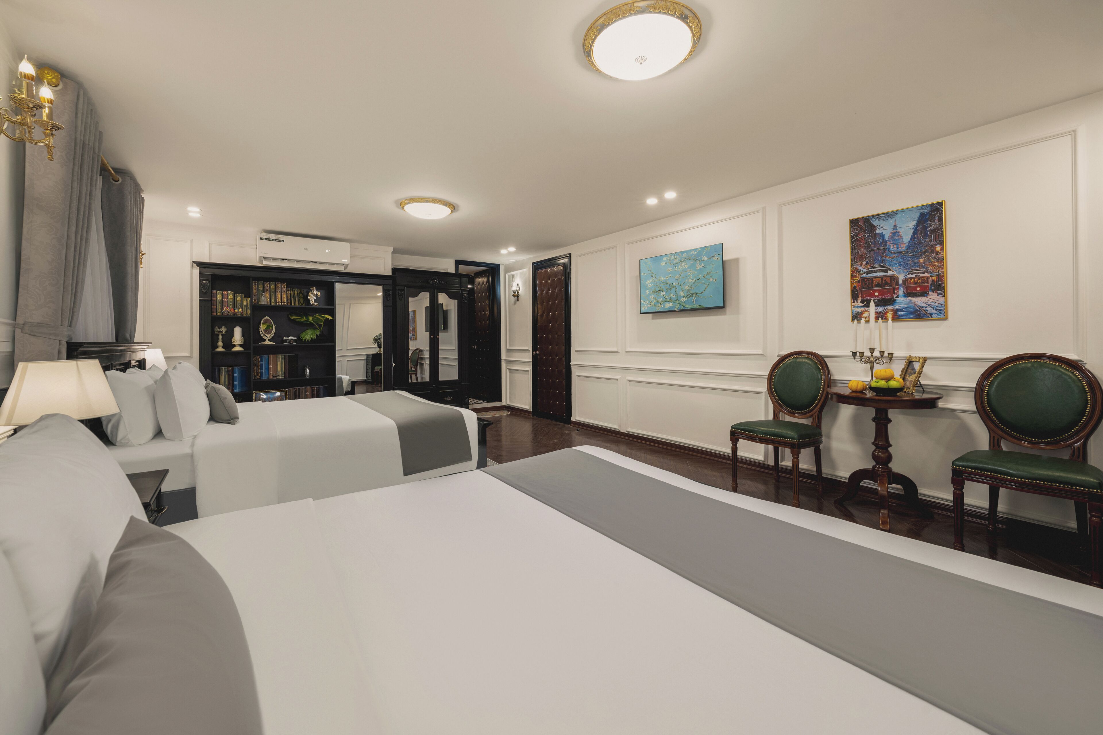 A hotel room with two beds, a desk with a chair, a bookshelf, and a painting on the wall.