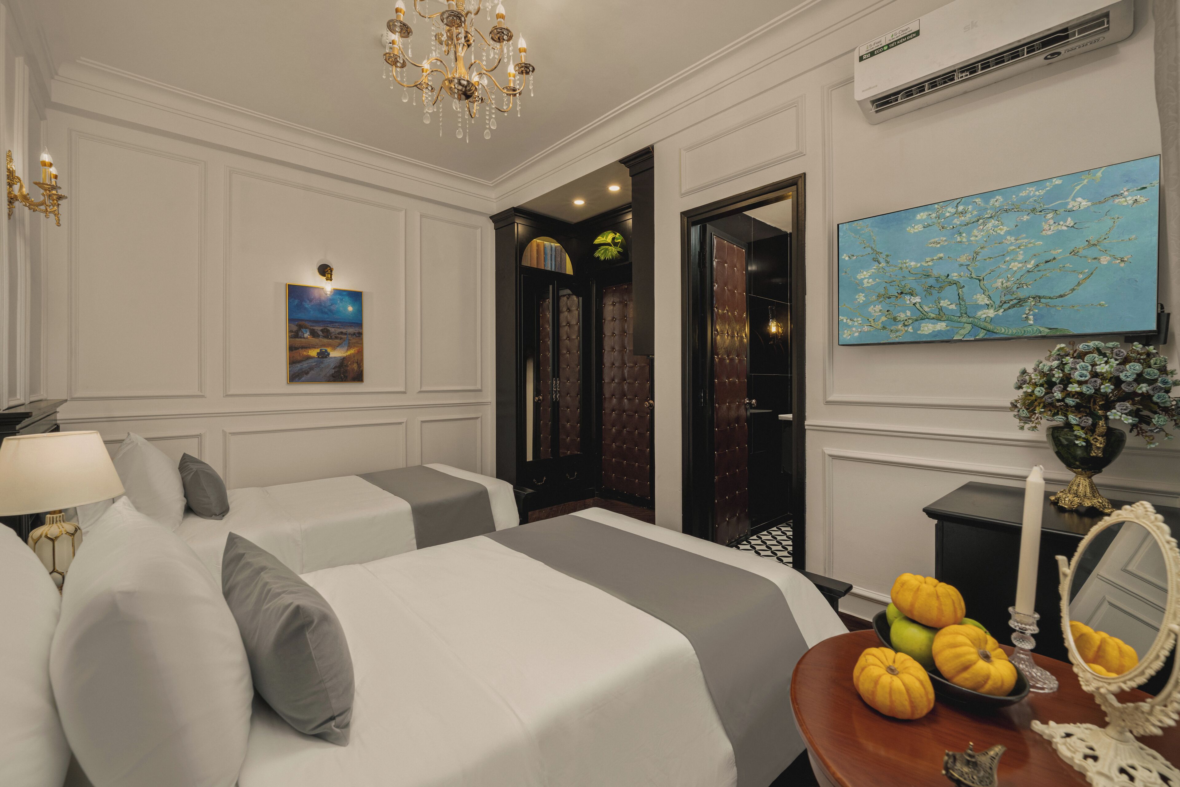 A hotel room with two beds, a chandelier, a painting, a mirror, and a vase with flowers.