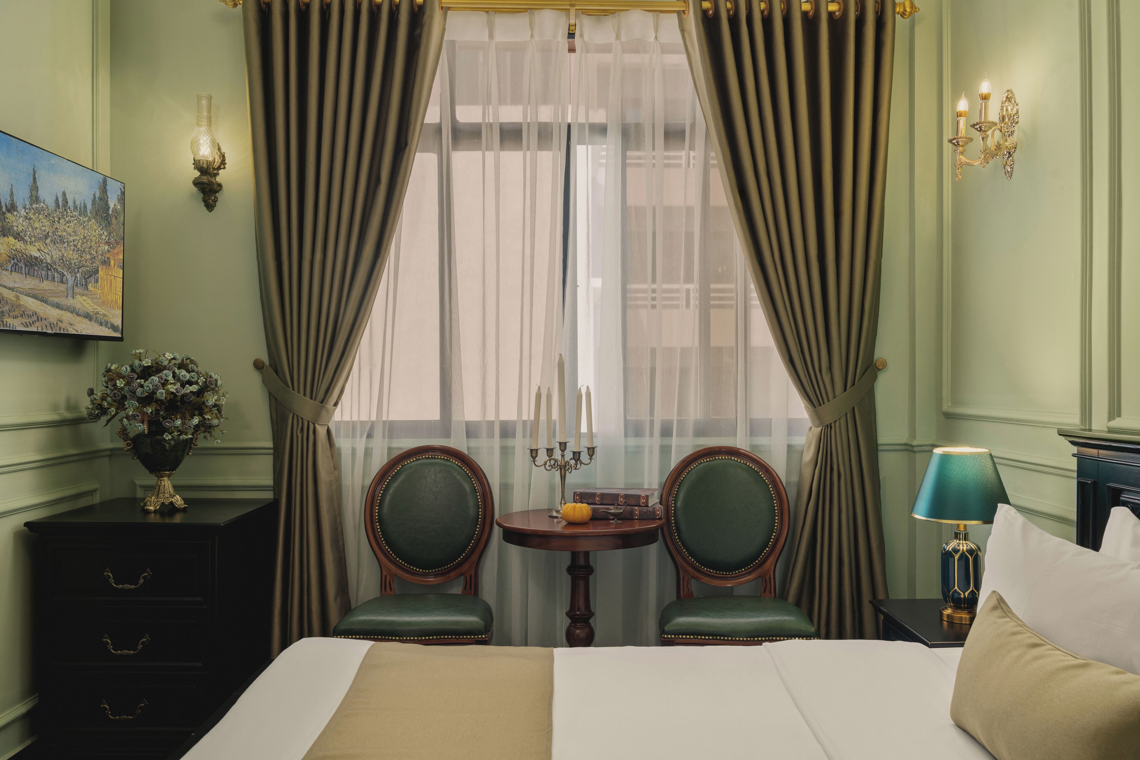 Hotelkamer met een bed, twee stoelen, een klein tafeltje met een bloevas, een lamp en een schilderij aan de muur.