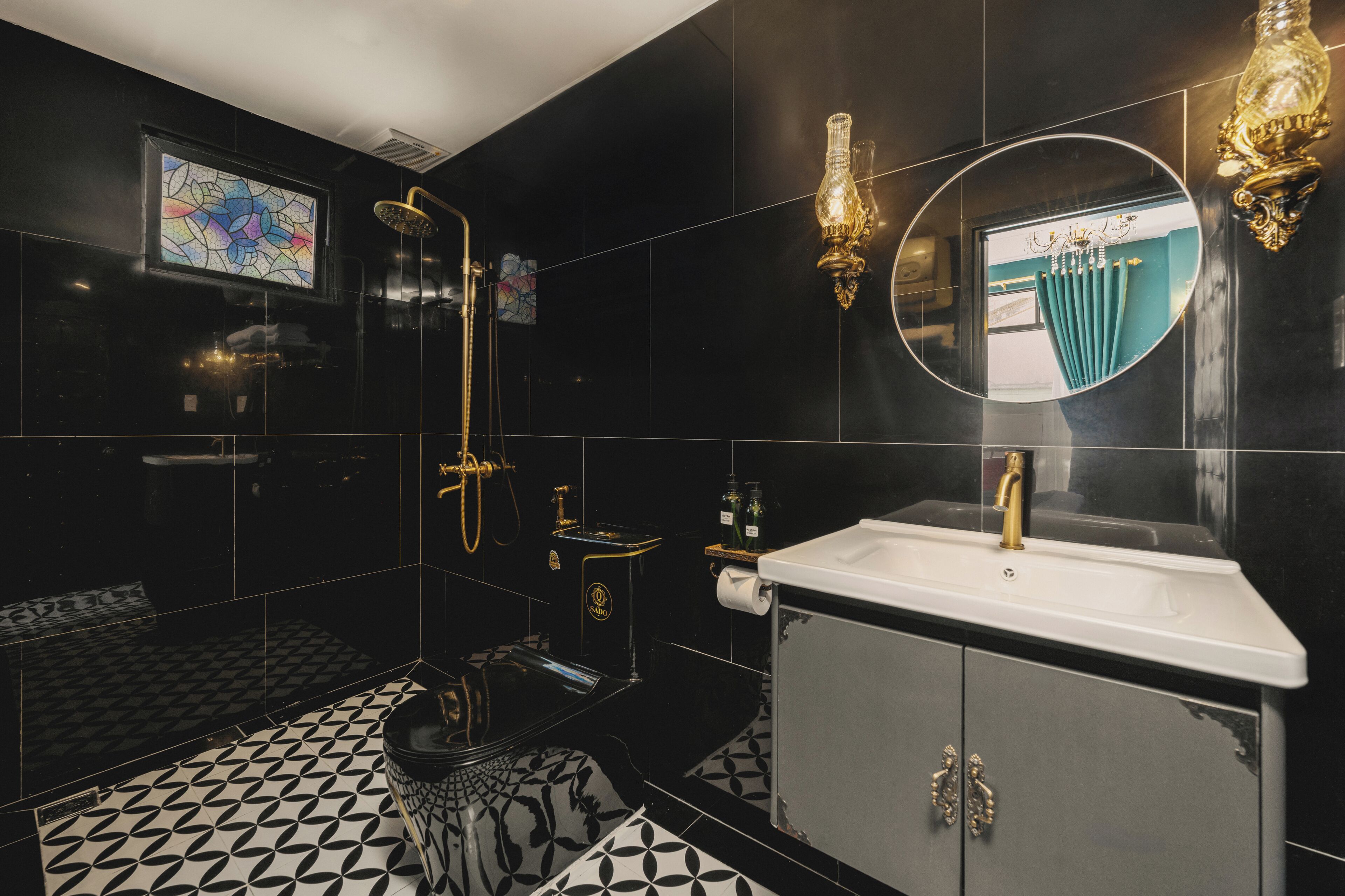 Un baño moderno con suelo de baldosas blanco y negro, un lavabo grande de color blanco con detalles dorados, una ducha con mampara de vidrio y un espejo decorativo con marco dorado.