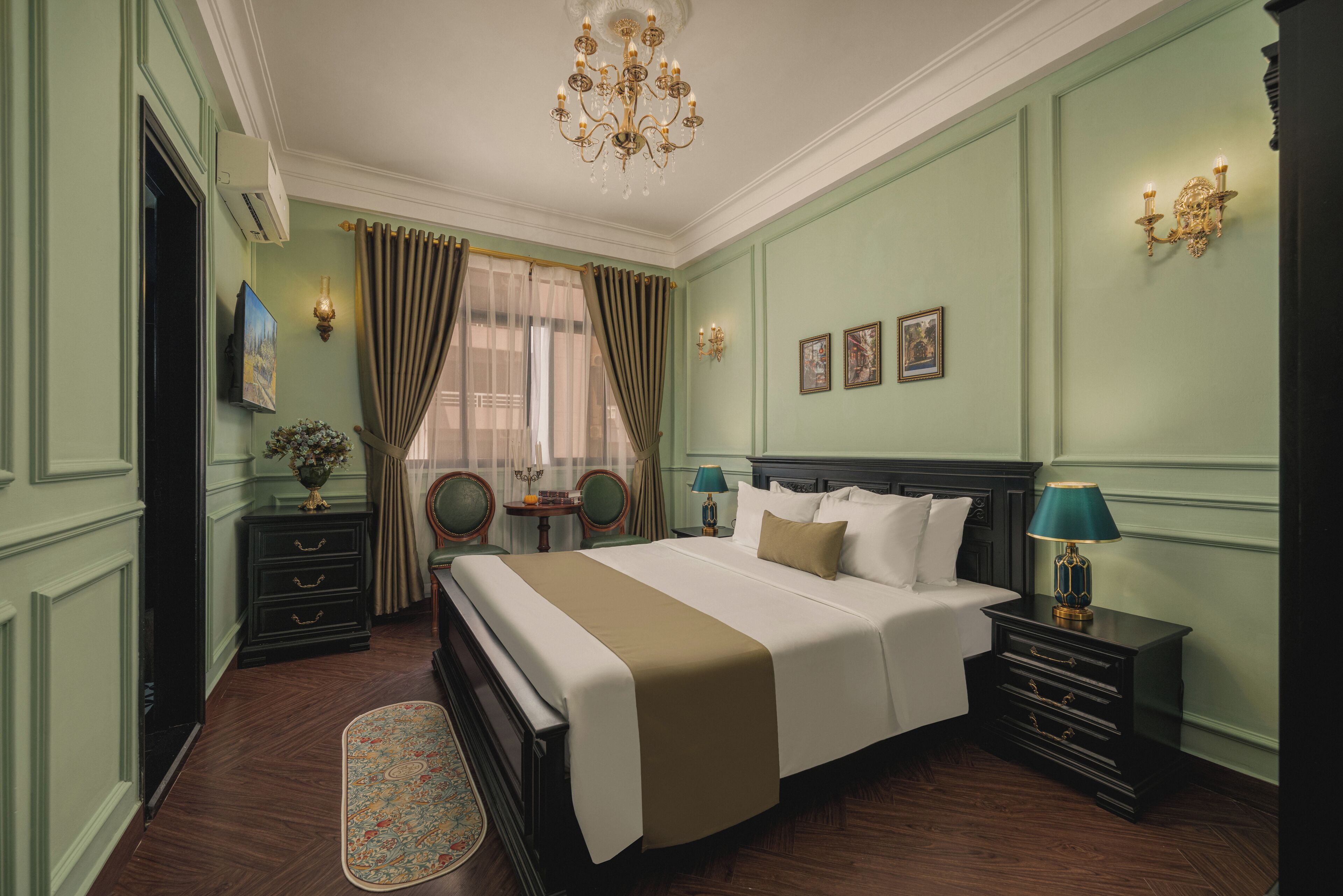 A hotel room with a large bed, a nightstand with a lamp, a dresser, a chair, and a desk with a vase of flowers.