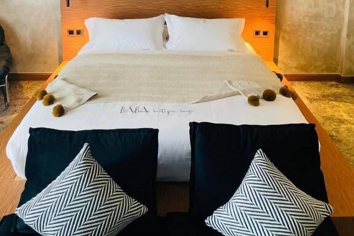 A modern bedroom with a wooden headboard, a bed with white linens, two black and white patterned pillows, and a bedside table.