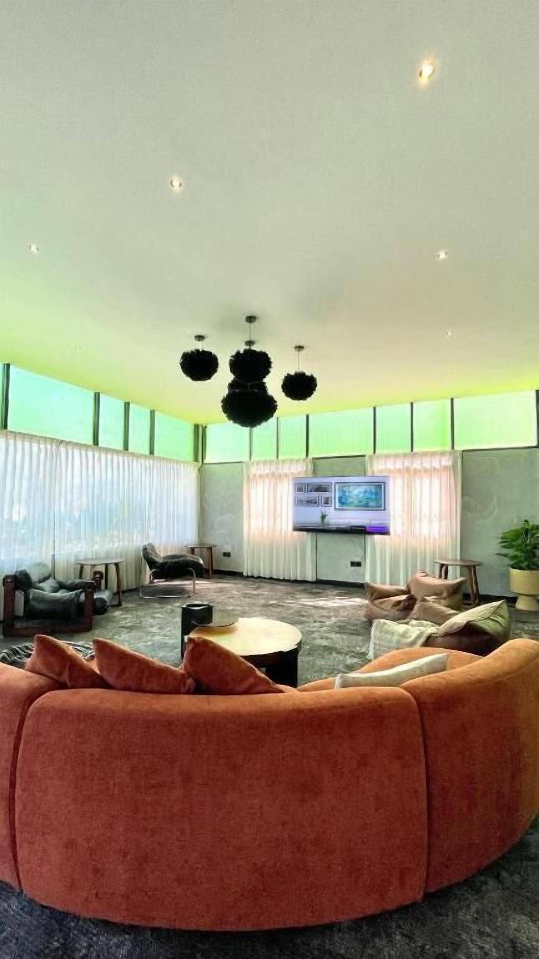 A modern living room with a large orange sofa, a flat-screen TV, and a large window with sheer curtains.
