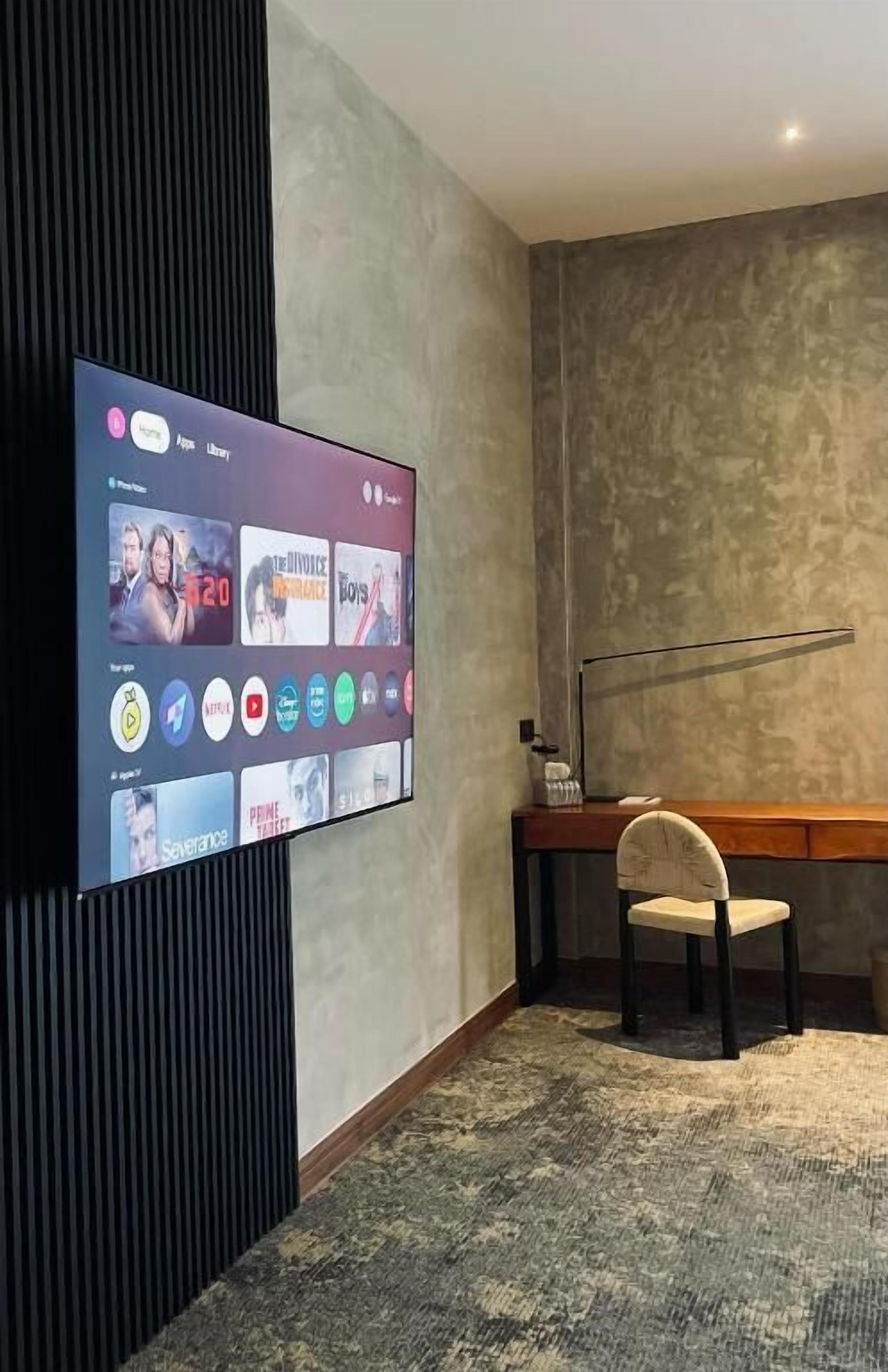 A modern room with a flat-screen TV displaying a streaming service interface, a desk with a lamp, and a chair.