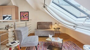 Ein gemütliches Schlafzimmer im Loft-Stil mit Dachfenster, einem Bett, einem Sessel, einem kleinen Tisch und einer Küchenzeile mit Kühlschrank und Backofen.