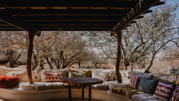A covered outdoor seating area with a curved sofa and a small table, surrounded by trees.