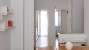 A modern, minimalist room with white walls, a wooden table, and a white bench. There is a large mirror on the wall and a hanging light fixture.