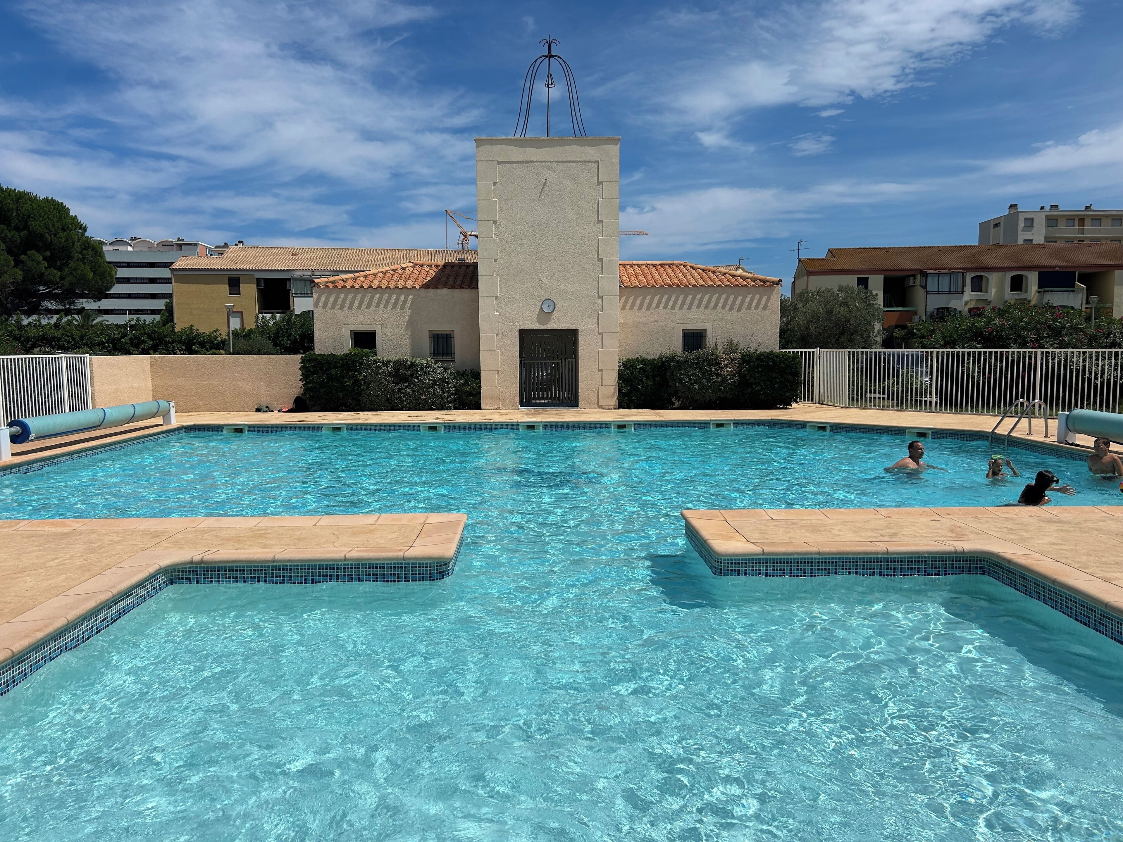 Ein rechteckiger Swimmingpool mit einer Fliesenleiste, umgeben von einem niedrigen Zaun, und ein Gebäude mit einem Flachdach im Hintergrund.