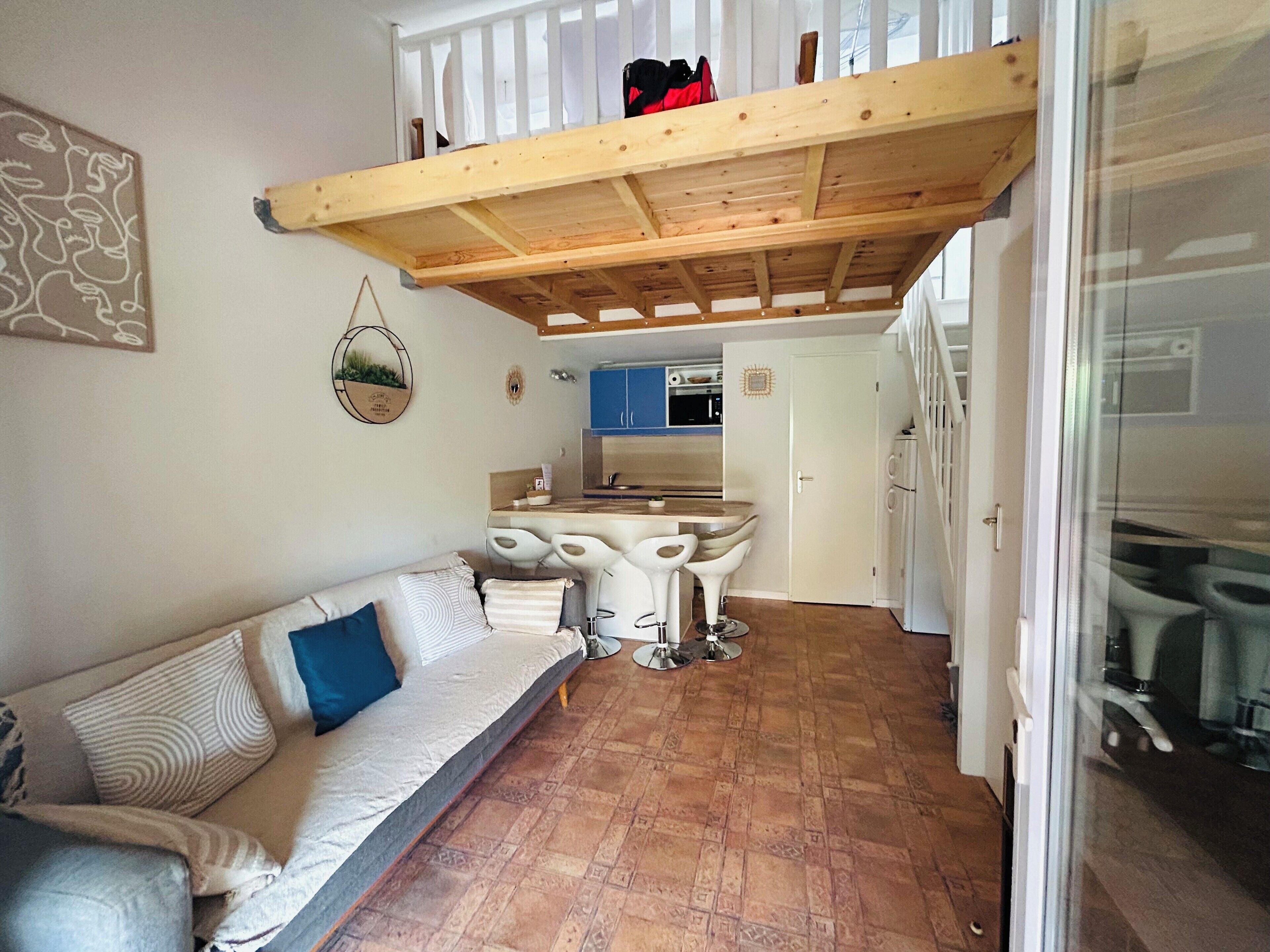 A cozy living area with a sofa, a dining table with four chairs, a kitchen in the background, and a wooden ceiling structure.