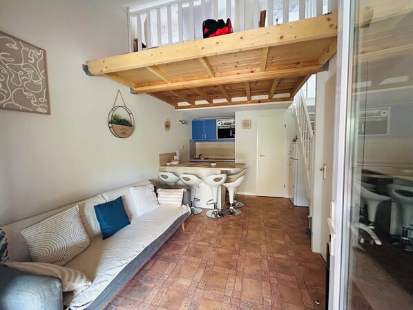 A cozy living area with a sofa, a dining table with four chairs, a kitchen in the background, and a wooden ceiling structure.