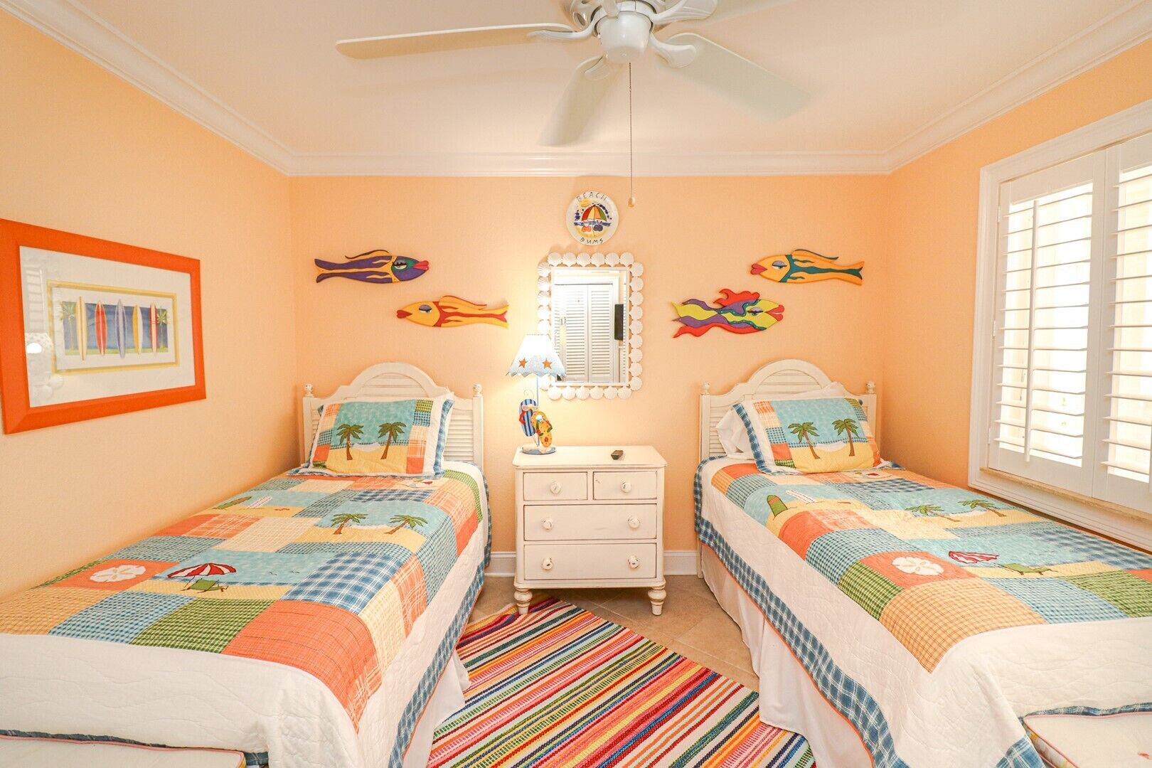 A bedroom with two beds, a ceiling fan, a window with shutters, and a colorful striped rug.