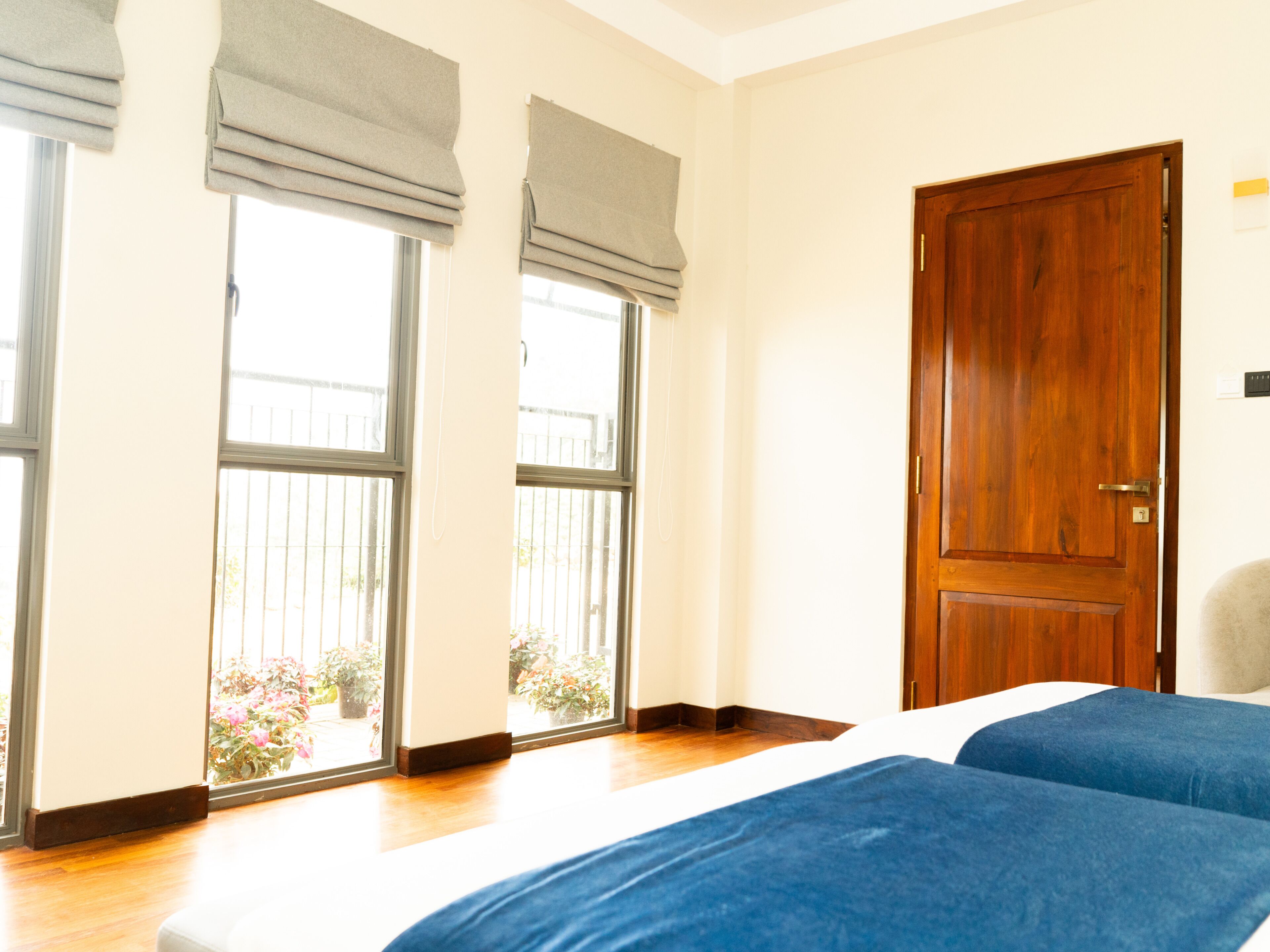 A hotel room with a wooden door, a large window with a view of plants, and a bed with blue bedding.