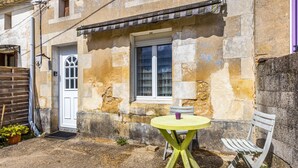 A stone building with a white door, a window with a metal grill, a small round table with a chair, and a wooden bench.