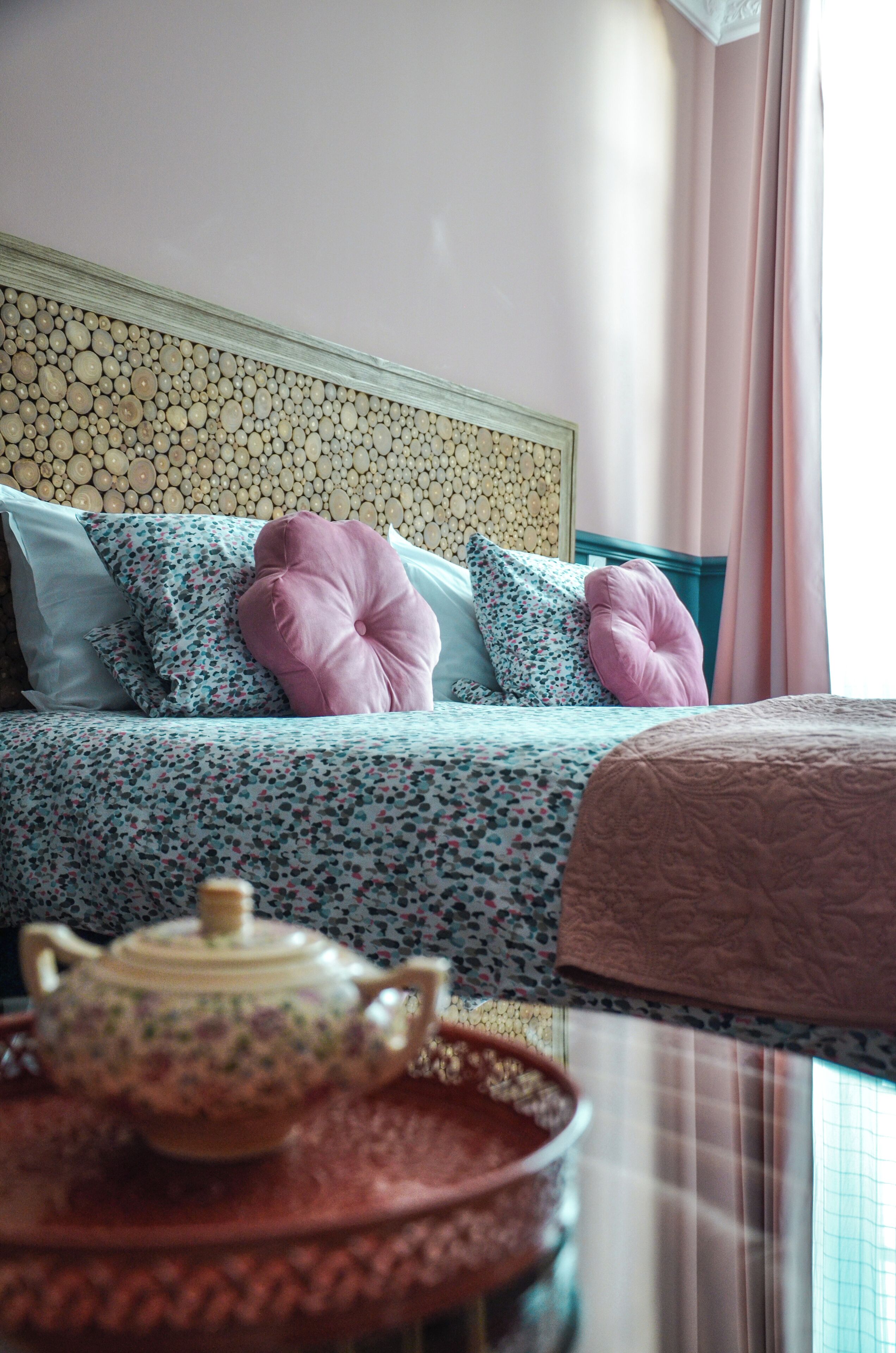 A neatly made bed with floral bedding and decorative pillows, a headboard with a patterned design, and a table with a teapot and tray in the foreground.