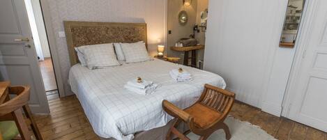 Un dormitorio ordenado con una cama grande, una silla de madera, una mesita de noche y un espejo en la pared.