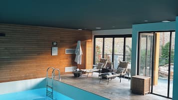 A modern indoor swimming pool with a wooden wall, lounge chairs, and a clear view of the outdoors.