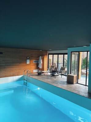 Una piscina interior moderna con pared de madera, sillones de descanso y vistas despejadas al exterior.