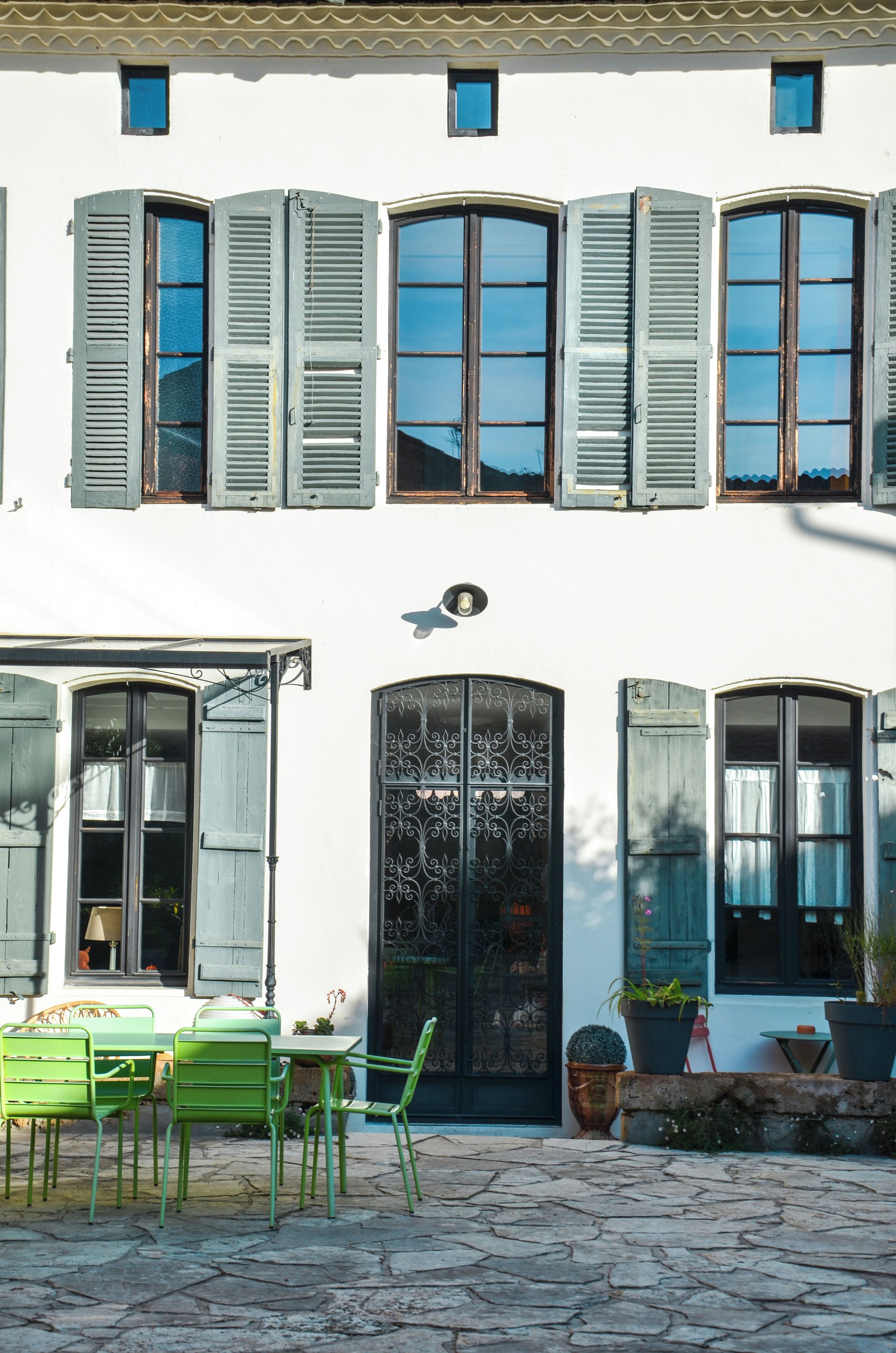 Un bâtiment à deux étages avec une façade blanche, de nombreuses fenêtres dotées de volets, une porte noire ornée et un patio pavé de pierres, agrémenté de chaises en métal vert et de plantes en pot.