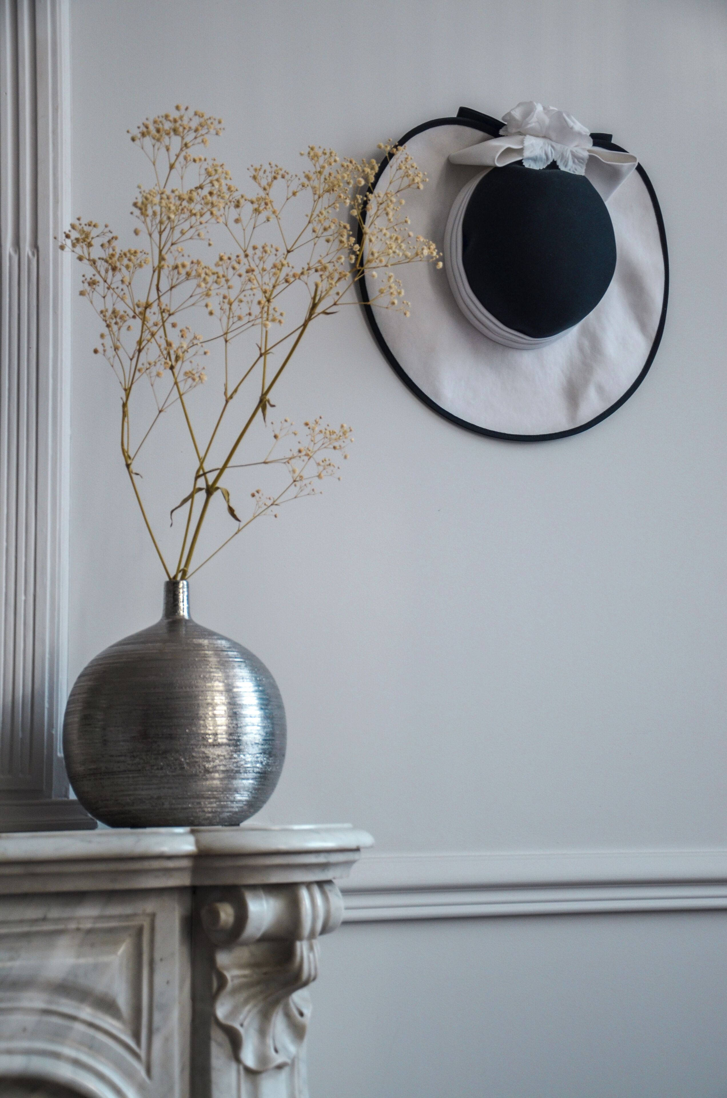 Um vaso com flores secas sobre um pedestal de mármore, um chapéu preto e branco com um laço na parede, e uma parede branca.