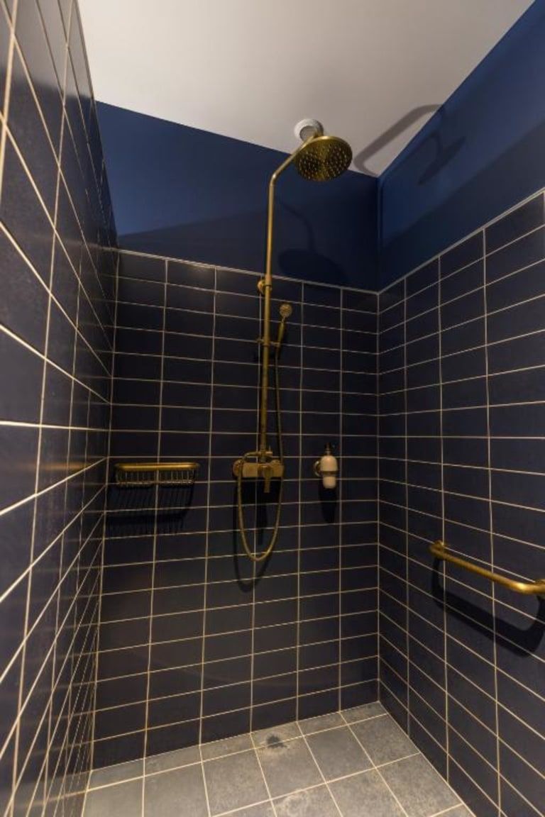Un baño pequeño revestido de azulejos azul oscuro, con ducha de cabeza dorada e hidromasaje manual.