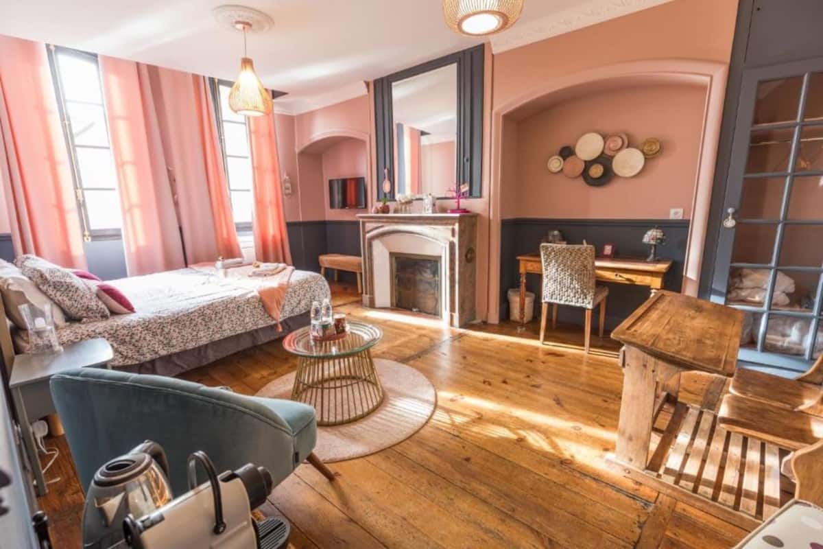 Una habitación acogedora con cama, zona de estar con sillón y mesa, chimenea y ventana con cortinas.