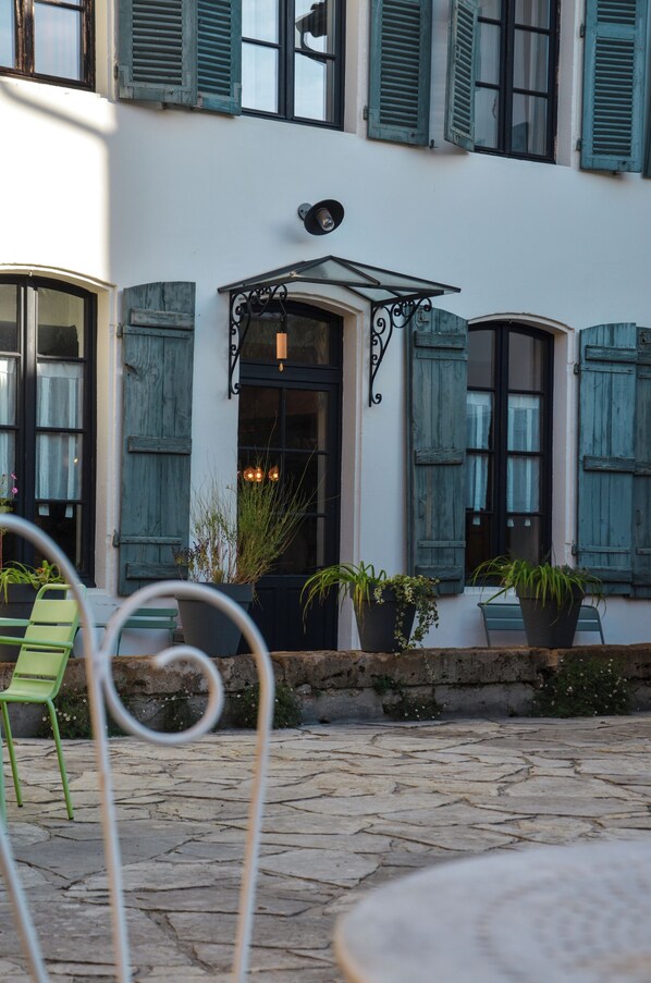 A building with a white facade, green shutters, and a stone-paved courtyard. There are potted plants and a green chair visible.
